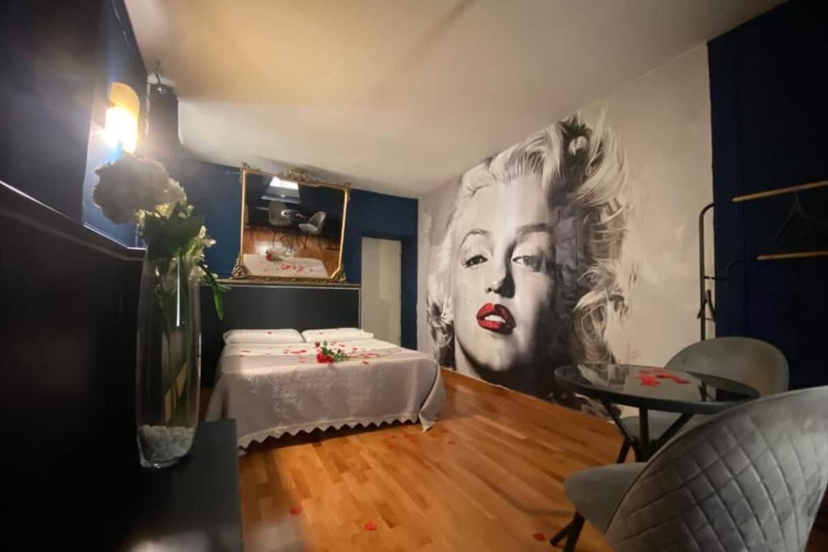 A room with a large mural, a bed with a white cover, a small table, and a chair.