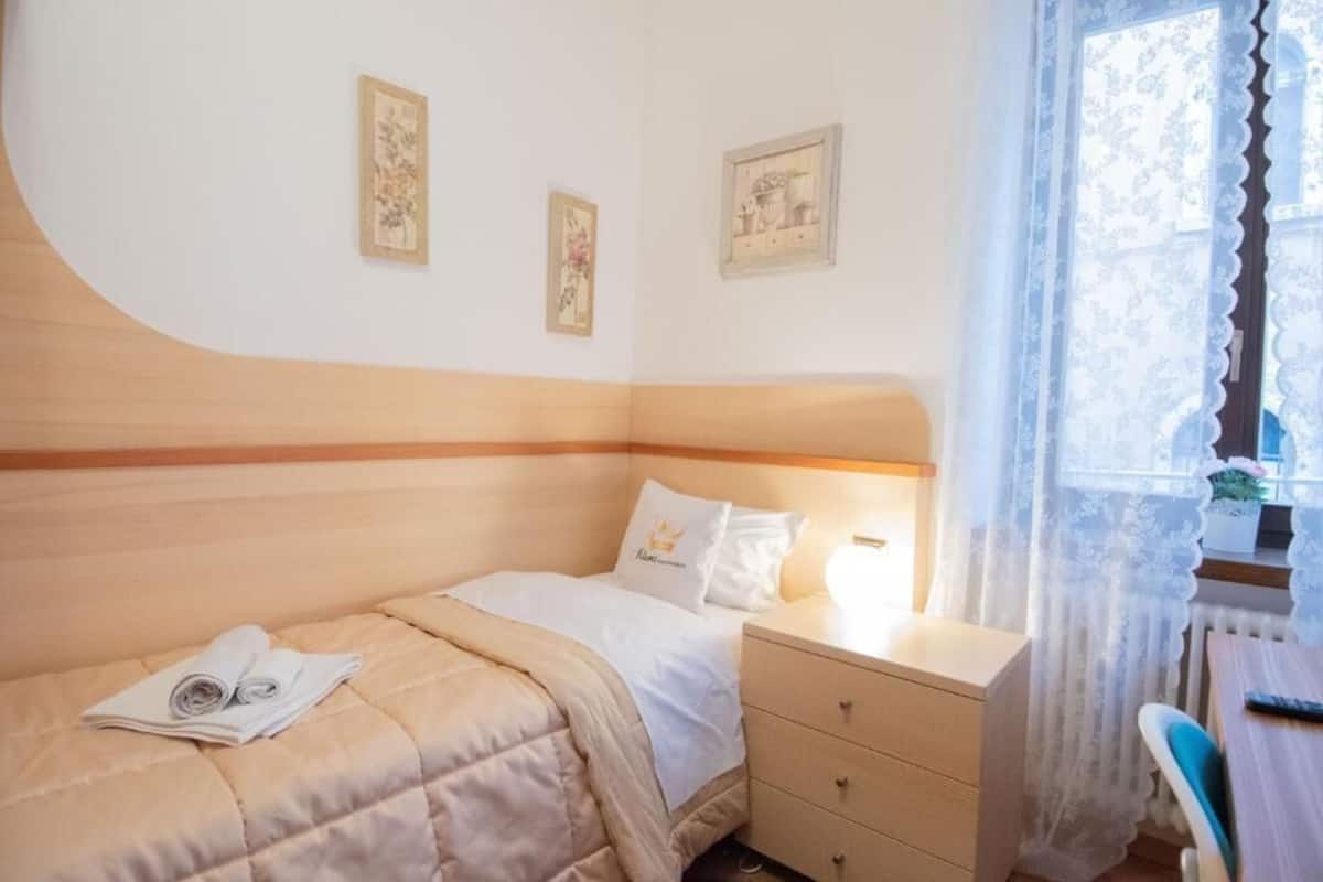 A bedroom with a bed, a bedside table, a chair, and a window with lace curtains.