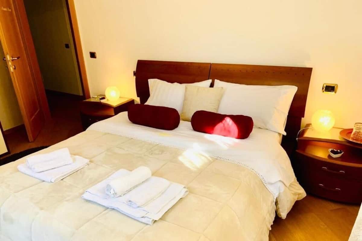 A neatly made bed with white and red pillows, a white duvet, and folded towels on the bed. There are two bedside tables with lamps and a wooden door in the background.
