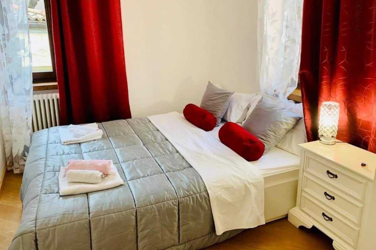 A neatly made bed with a grey quilt, white linens, and red pillows. There is a white bedside table with drawers and a lamp.