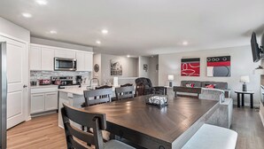 A modern kitchen with white cabinets, a central island, and a dining area with a large wooden table and grey upholstered chairs. - The Magnolia Retreat (Omaha)