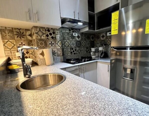 Una cocina con un lavamanos de acero inoxidable, encimera de granito y un refrigerador.