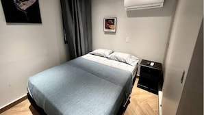 Un dormitorio con una cama, una mesita de noche, aire acondicionado y un cuadro en la pared.
