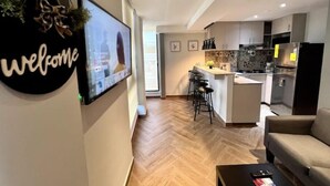 Una sala de estar moderna con un sofá y una mesa de centro, una cocina con barra y un televisor en la pared mostrando un mensaje de bienvenida.