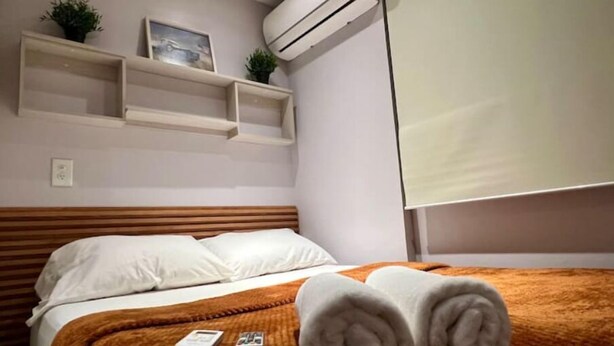 A hotel room with a bed, white pillows, a brown blanket, and a rolled towel on the bed. There is an air conditioning unit on the wall and a small shelf with a plant and a picture frame.