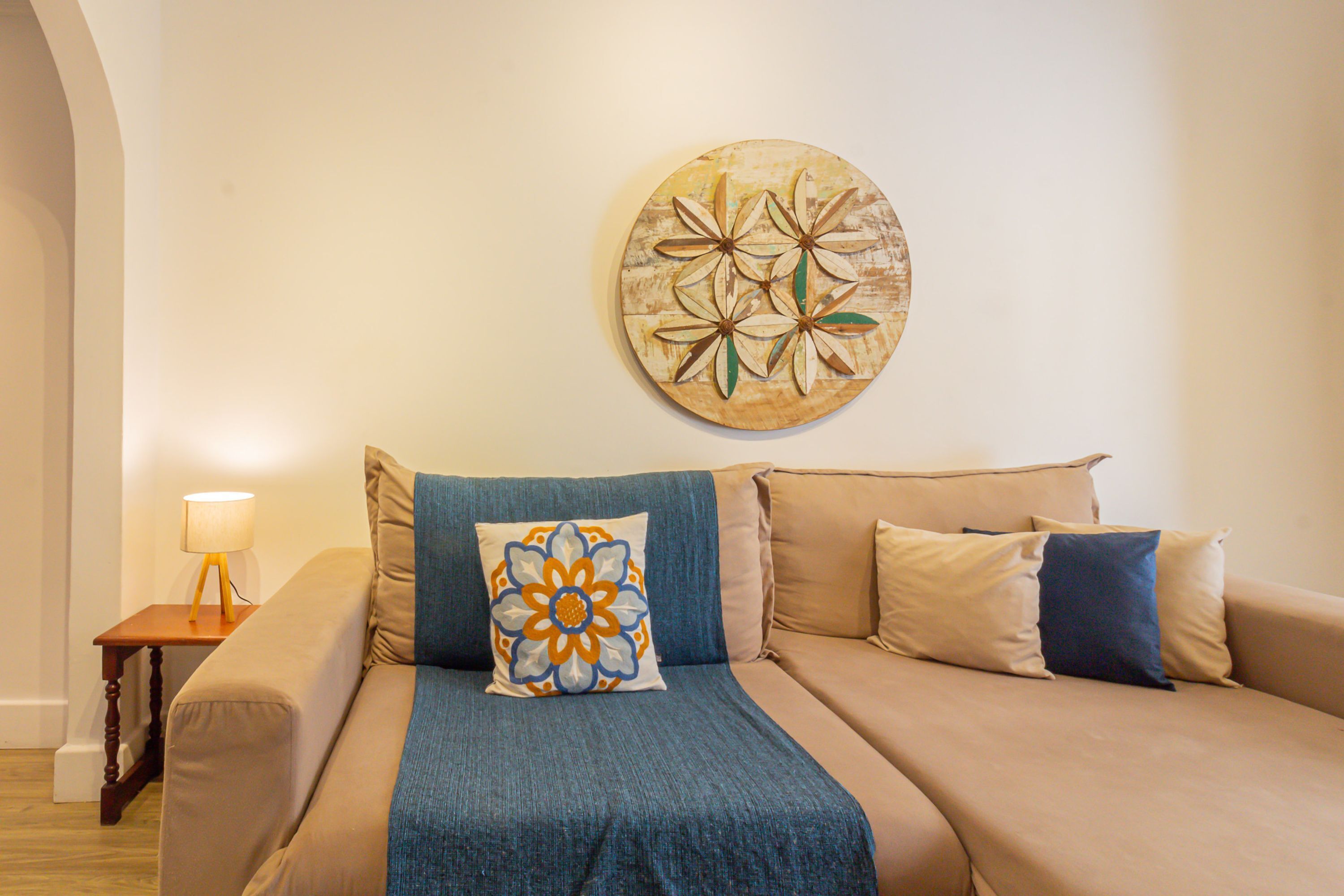 A beige sofa with a blue throw blanket and decorative pillows, a wooden side table with a lamp, and a wall-mounted circular artwork.