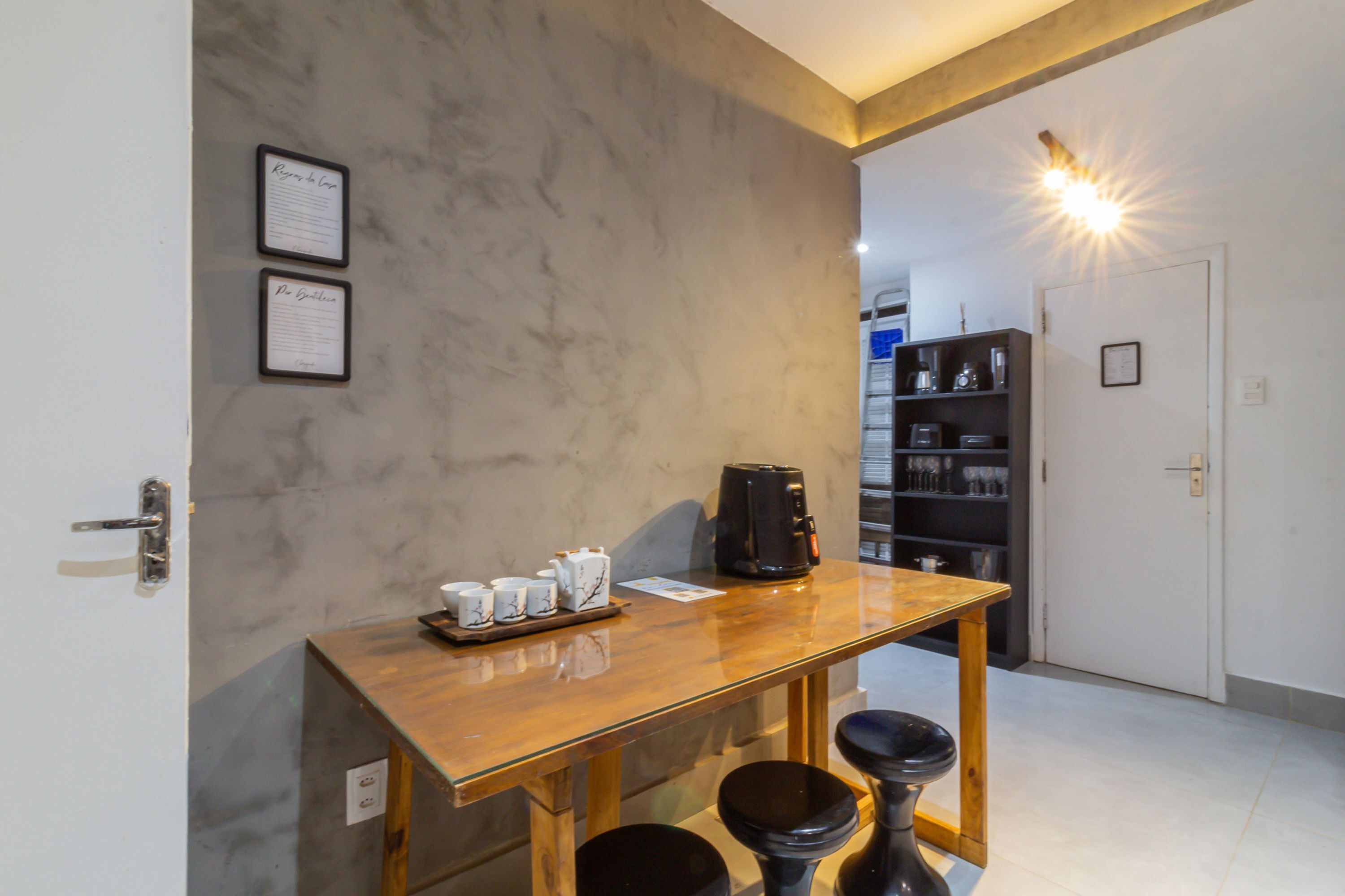 A small kitchen area with a wooden table, two bar stools, a coffee maker, and a shelf with kitchen items.