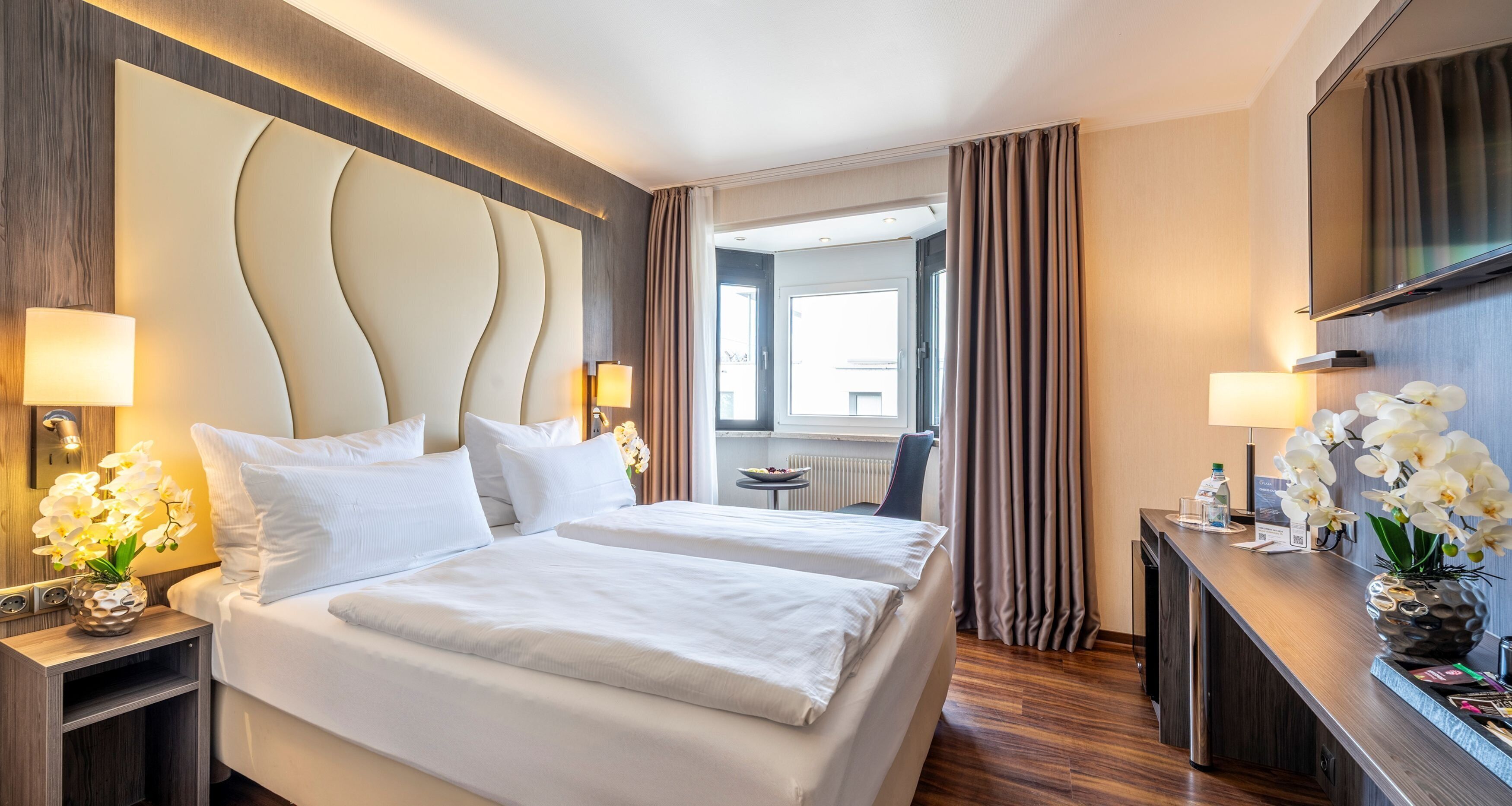 Een hotelkamer met een groot bed, twee nachtkastjes met lampen, een aan de muur bevestigde televisie en een bureau met een bloemenvaas.