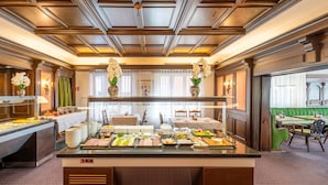 Buffet com diversos alimentos, área de estar com mesas e cadeiras, e teto decorativo.
