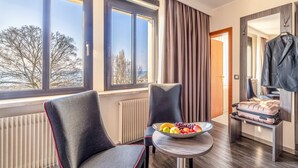 Quarto de hotel com uma janela grande, um radiador, uma mesa com uma fruteira, duas cadeiras, um cabideiro com um casaco e uma mala num armário.