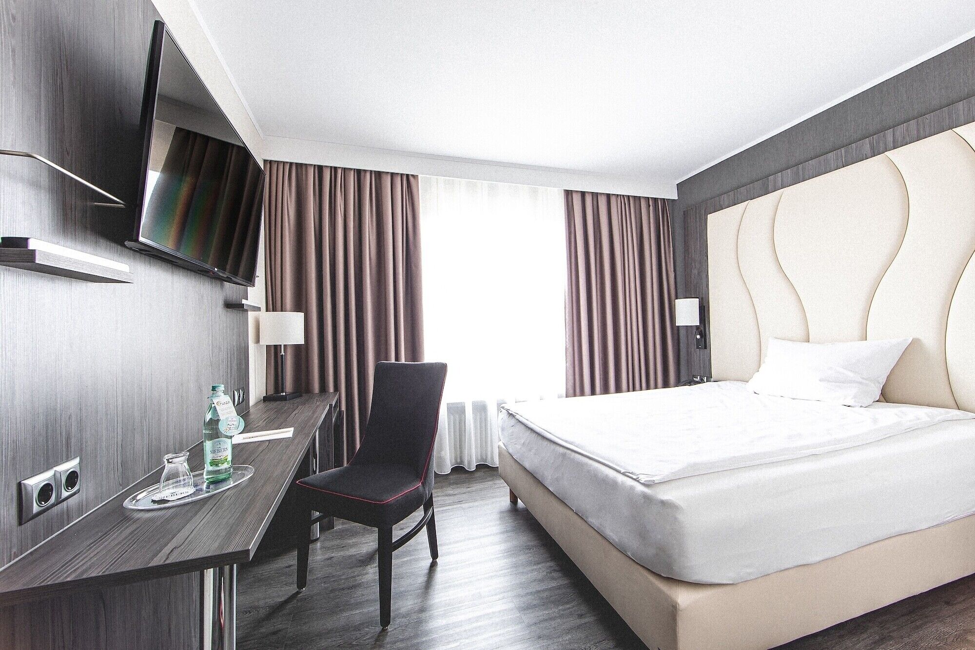 A hotel room with a bed, a desk, a chair, a TV, and curtains.