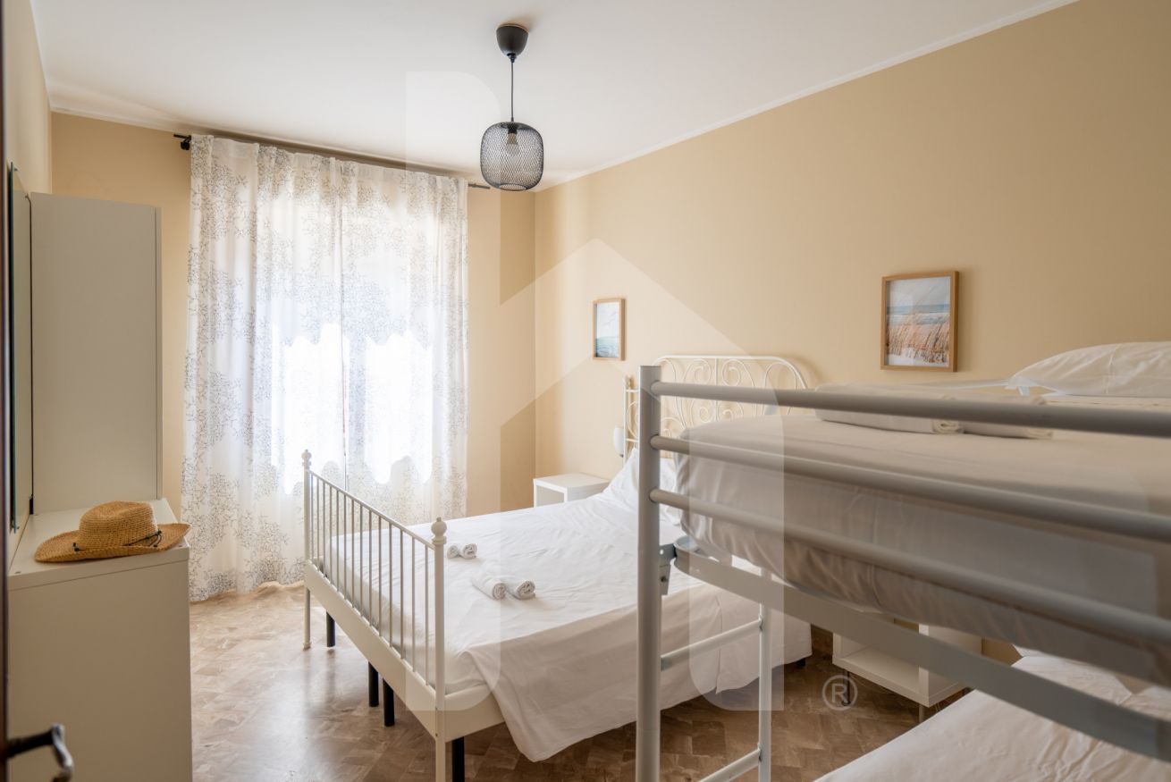A hotel room with a bunk bed, a single bed, a small bedside table, a wardrobe, and a window with sheer curtains.
