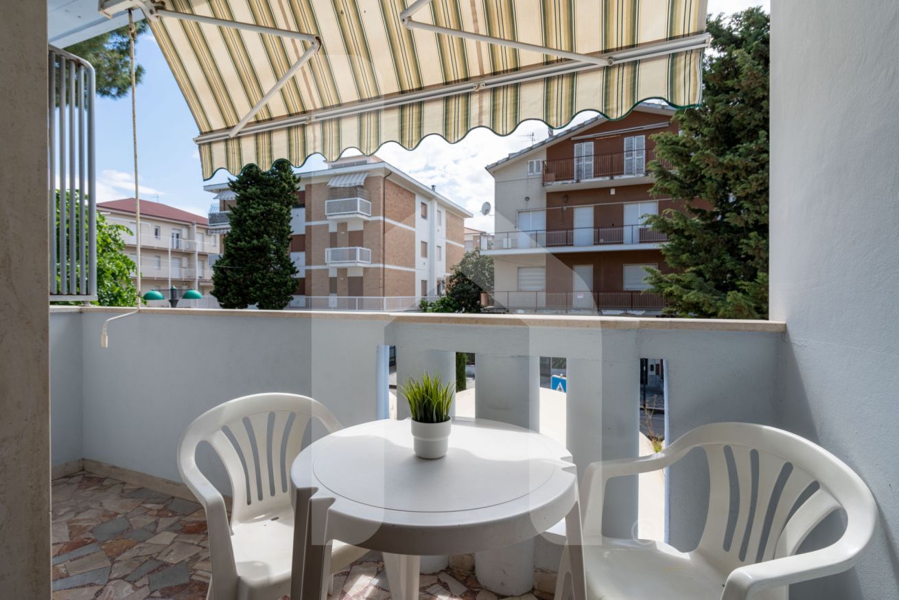 Un balcón exterior con una mesa blanca y dos sillas blancas, un toldo a rayas y vistas a edificios residenciales.