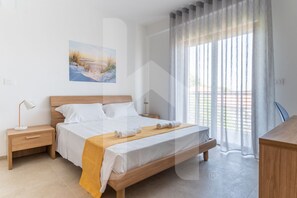 A modern bedroom with a wooden bed, white linens, a yellow throw, a nightstand with a lamp, and a painting on the wall.