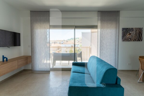 A modern living room with a teal sofa, a flat-screen TV, a wooden console, and a sliding glass door leading to a balcony with outdoor furniture.