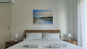 Een opgemaakt bed met witte lakens en twee kussens, een aan de muur hangend schilderij van een strandgezicht, en een raam met witte gordijnen.