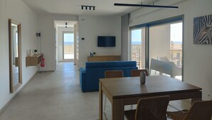Een moderne woonkamer met een bank, een eettafel en een televisie. Er is een balkon met uitzicht op zee.