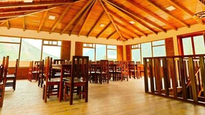 A spacious hall with wooden floors and a high ceiling, featuring rows of wooden chairs and tables, large windows, and a mountainous view.