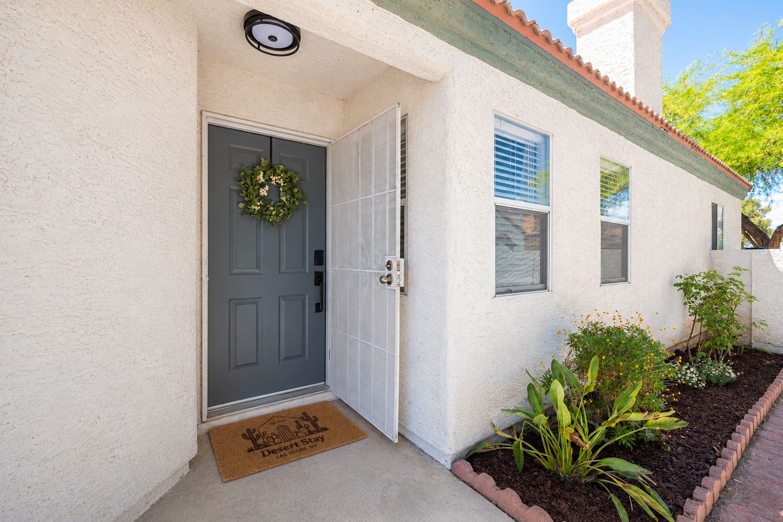 A house with a grey door, a doormat, and a small garden with plants and a brick border.
