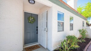 Entrada de una casa con puerta gris, un felpudo que dice 'HELLO' y un pequeño jardín con plantas y flores.