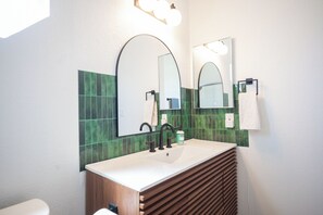 Ein modernes Badezimmer mit Waschbecken in Weiß, zwei Spiegeln, einer grün gefliesten Rückwand und einer Holz-Waschtischunterschrank.