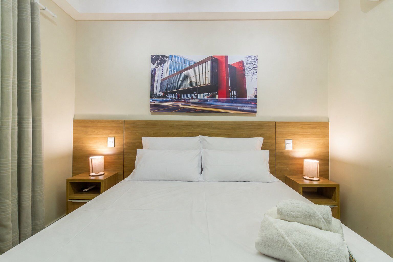 Habitación de hotel con una cama, dos mesitas de noche con lámparas, un cuadro en la pared y una ventana con cortinas.