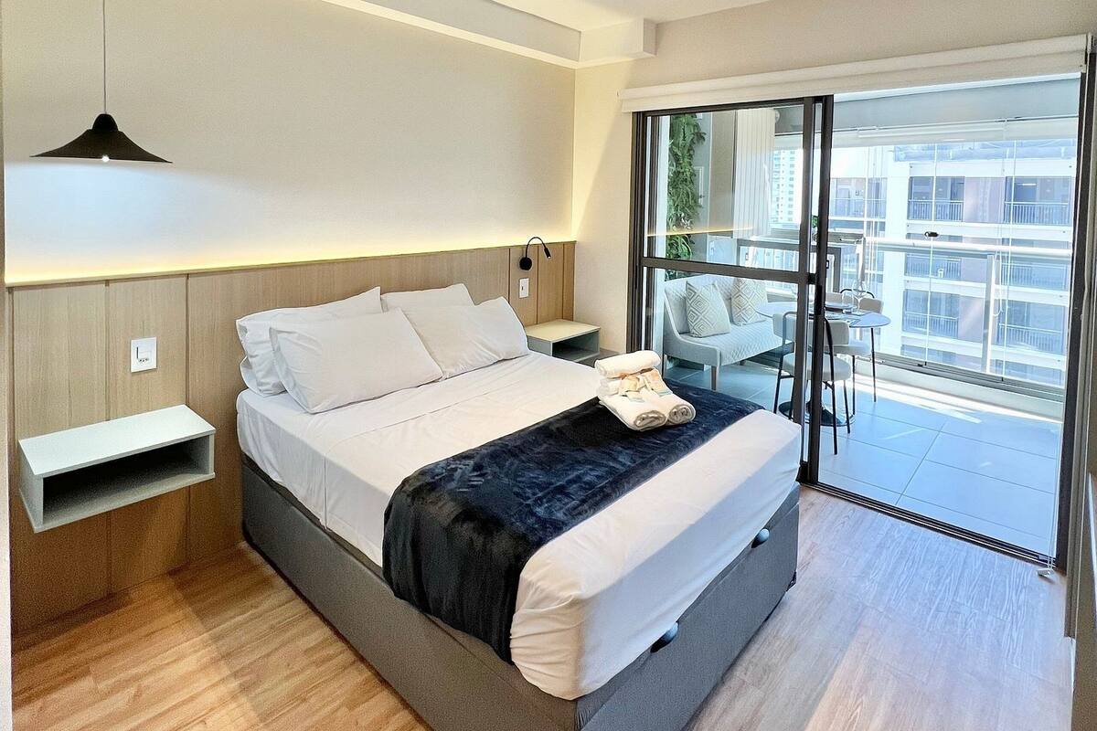 Moderni hotellihuone, jossa on suuri sänky, yöpöydät, pieni työpöytä ja parveke, jolta on näkymä.