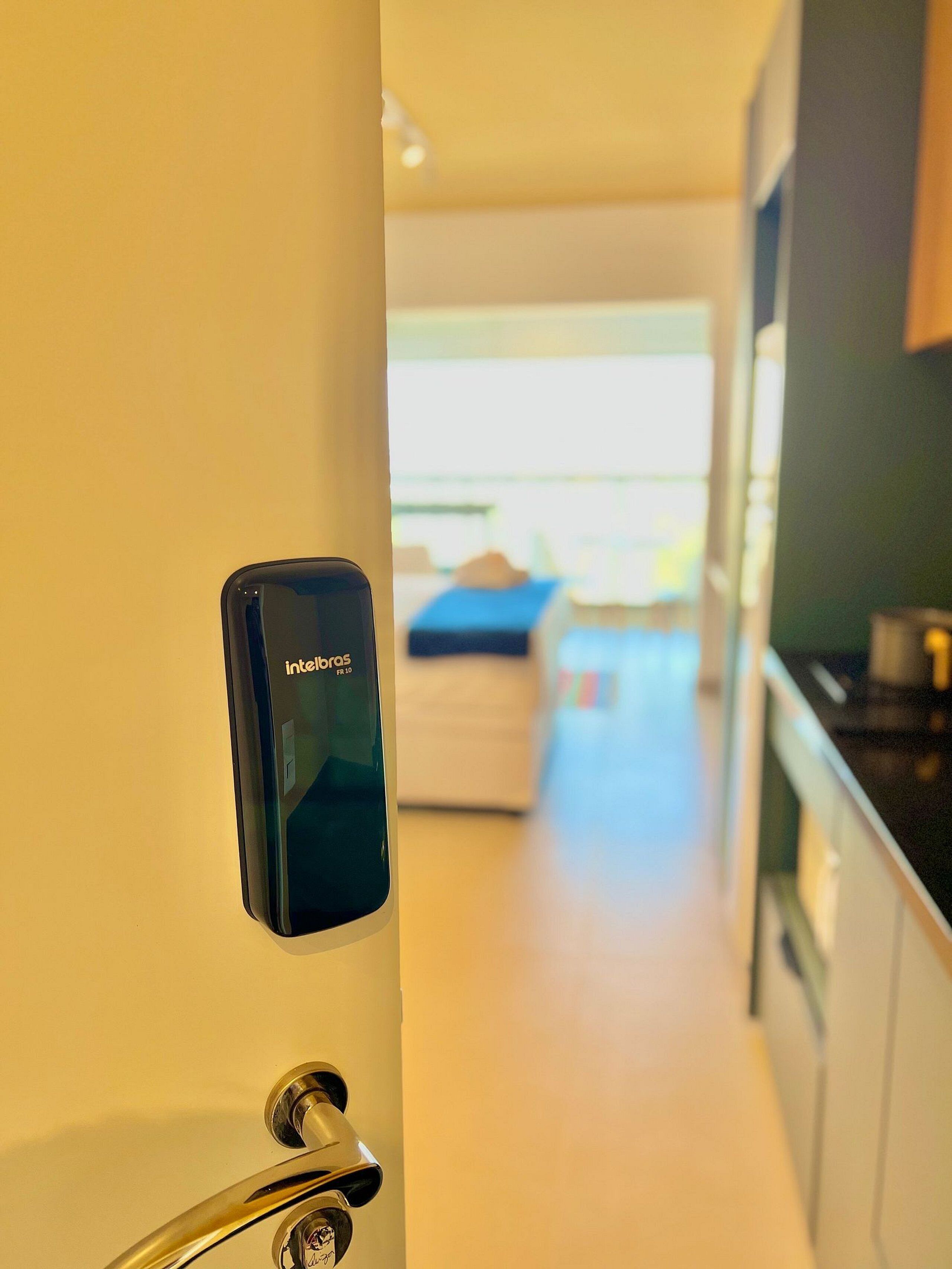 Una cocina moderna con cerradura digital, un ventanal visible y un vistazo a un dormitorio con cama y manta azul.