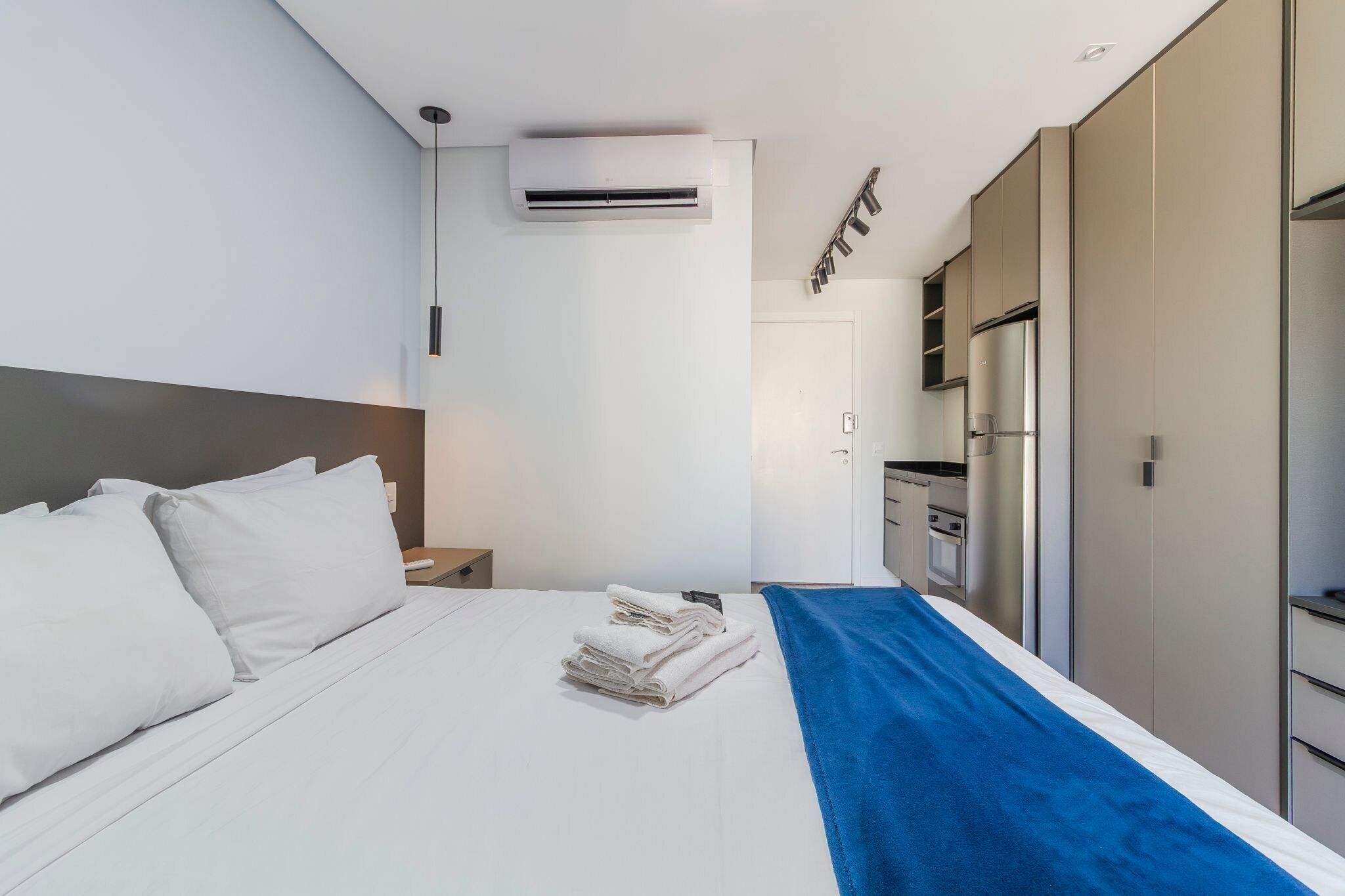A modern hotel room with a neatly made bed, an air conditioning unit, a small kitchen area, and a wardrobe.