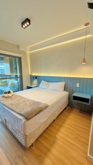 Une chambre d’hôtel moderne avec un grand lit, des tables de chevet, une petite table, une fenêtre avec des stores et un luminaire suspendu.