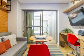 Una sala moderna con un sofá, una mesa de comedor para dos personas, una silla roja, un televisor y un balcón al que se accede por una puerta corrediza.