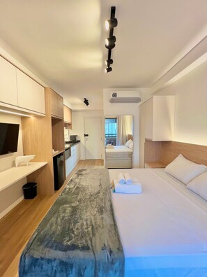 Habitación de hotel con cama, una banca, escritorio, televisor, zona de cocina y aire acondicionado.