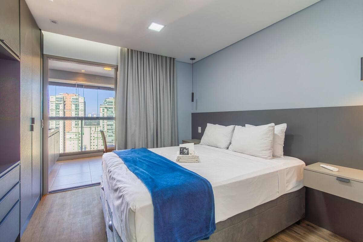 Hotellihuone, jossa on sänky, yöpöytä, ikkuna verhoilla ja näkymä rakennuksiin.