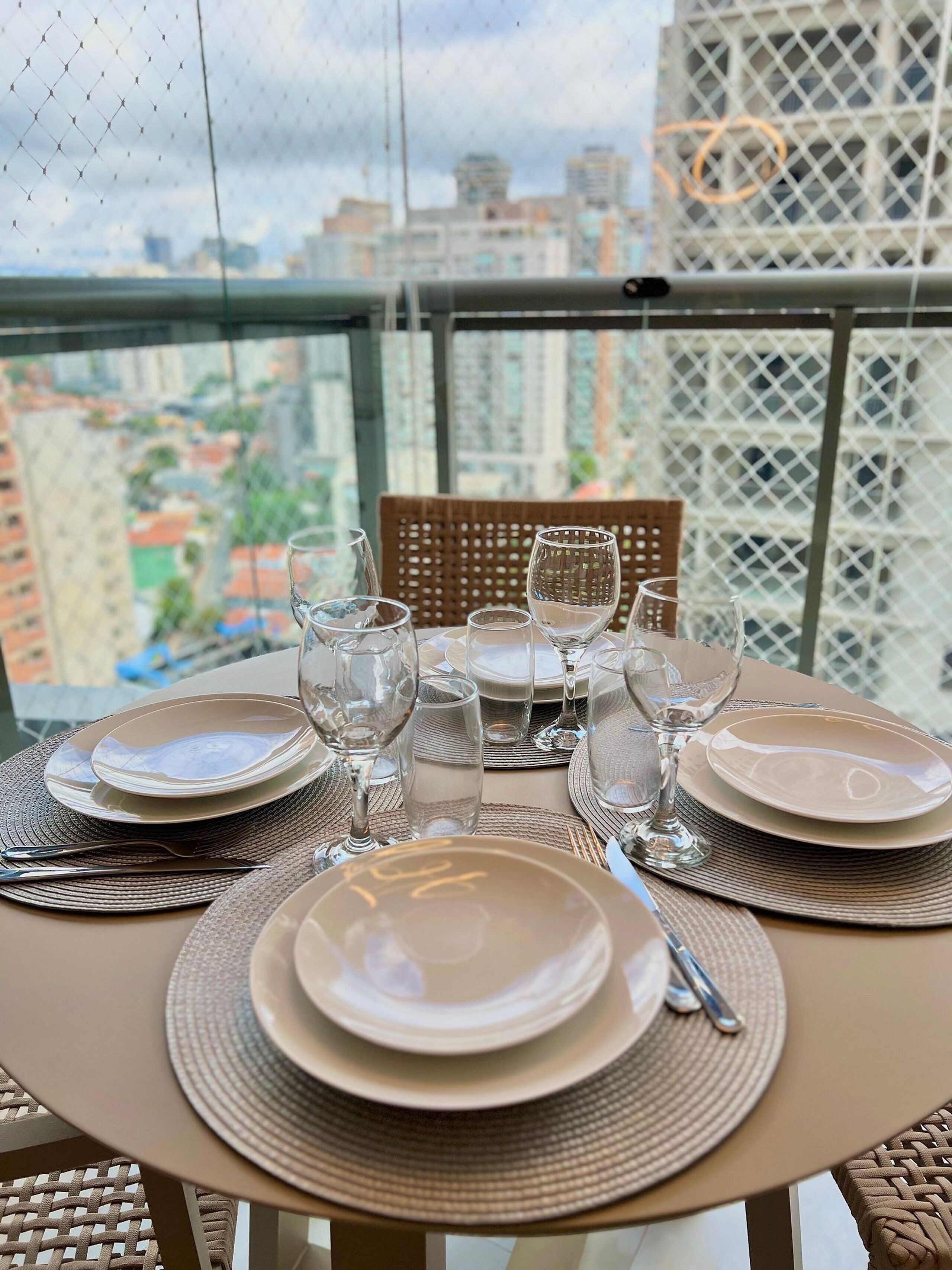 A table set for dining with clear glassware and plates, a wicker chair, and a view of a cityscape through a mesh window.