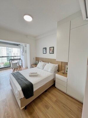Habitación de hotel moderna con una cama grande, una mesita de noche, un armario y un balcón con vistas a la ciudad.