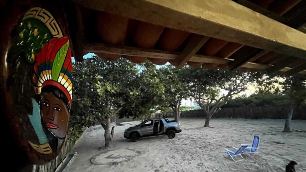 Un espacio al aire libre cubierto con un coche estacionado bajo una estructura de madera, árboles y una silla de playa.