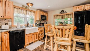 A kitchen with wooden cabinets, a black refrigerator, a microwave, a toaster, and a dining area with wooden chairs. - Cozy Lil Red Cabin; Water Frontage, Dog Friendly! (Ellsworth)