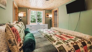 A room with a wooden ceiling, a bed with a quilt, a dresser, a window with curtains, and a mounted TV. - Cozy Lil Red Cabin; Water Frontage, Dog Friendly! (Ellsworth)