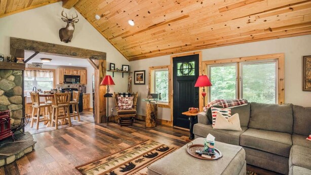 A rustic interior with a wooden ceiling, a stone wall, a dining area, and a cozy living space with a sofa and a fireplace. - Cozy Lil Red Cabin; Water Frontage, Dog Friendly! (Ellsworth)
