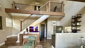 Un vaste espace de vie agrémenté d’un escalier en bois, d’un fauteuil vert, d’un téléviseur à écran plat et d’un coin cuisine doté d’un plan de travail blanc.