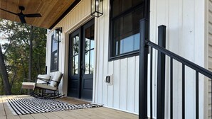 Une véranda en bois avec une chaise berçante et un tapis, une porte noire et une rambarde noire.