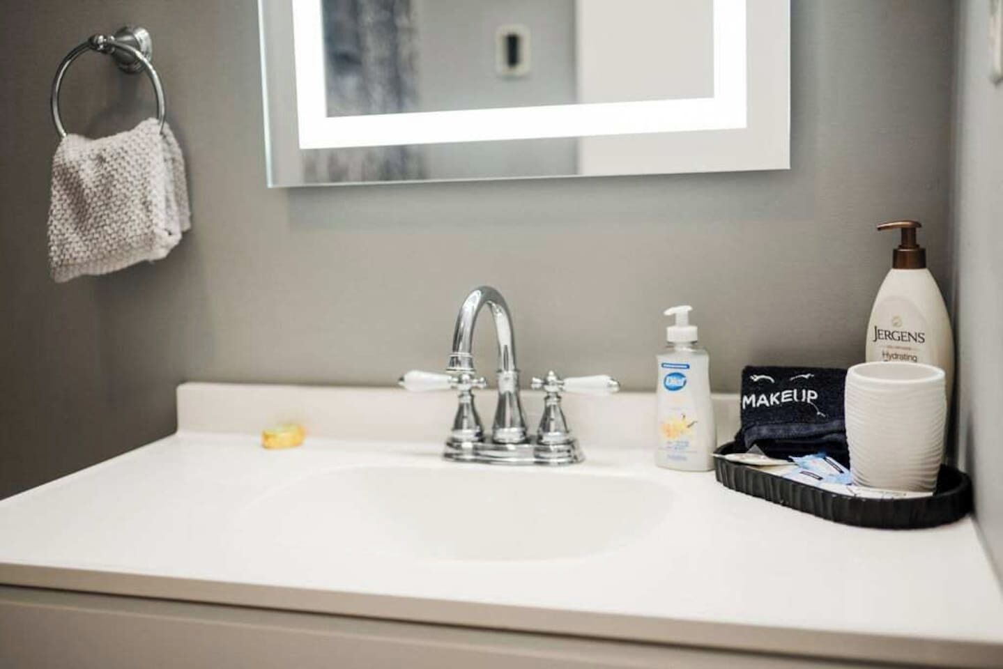 A bathroom with a white sink, a mirror, a towel rack, a soap dispenser, a Jergens lotion bottle, and a makeup bag.