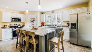 Una cocina con gabinetes blancos, electrodomésticos de acero inoxidable, una isla central con taburetes altos e iluminación colgante.