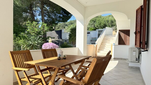 Une terrasse extérieure avec une table et des chaises en bois, une vue sur un escalier et un jardin avec des arbres et des fleurs.