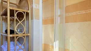 Une salle de bain dotée d’une douche à parois de verre, d’une étagère en bambou où sont posées des serviettes et des articles de toilette, et d’un mur carrelé agrémenté d’une bordure décorative.