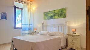 Une chambre à coucher comprenant un lit, deux tables de chevet, une fenêtre avec des rideaux, un tableau accroché au mur et une petite table de nuit avec une lampe.