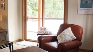 A living room with a brown leather armchair, a matching ottoman, a glass coffee table, and a large sliding glass door leading to a deck.