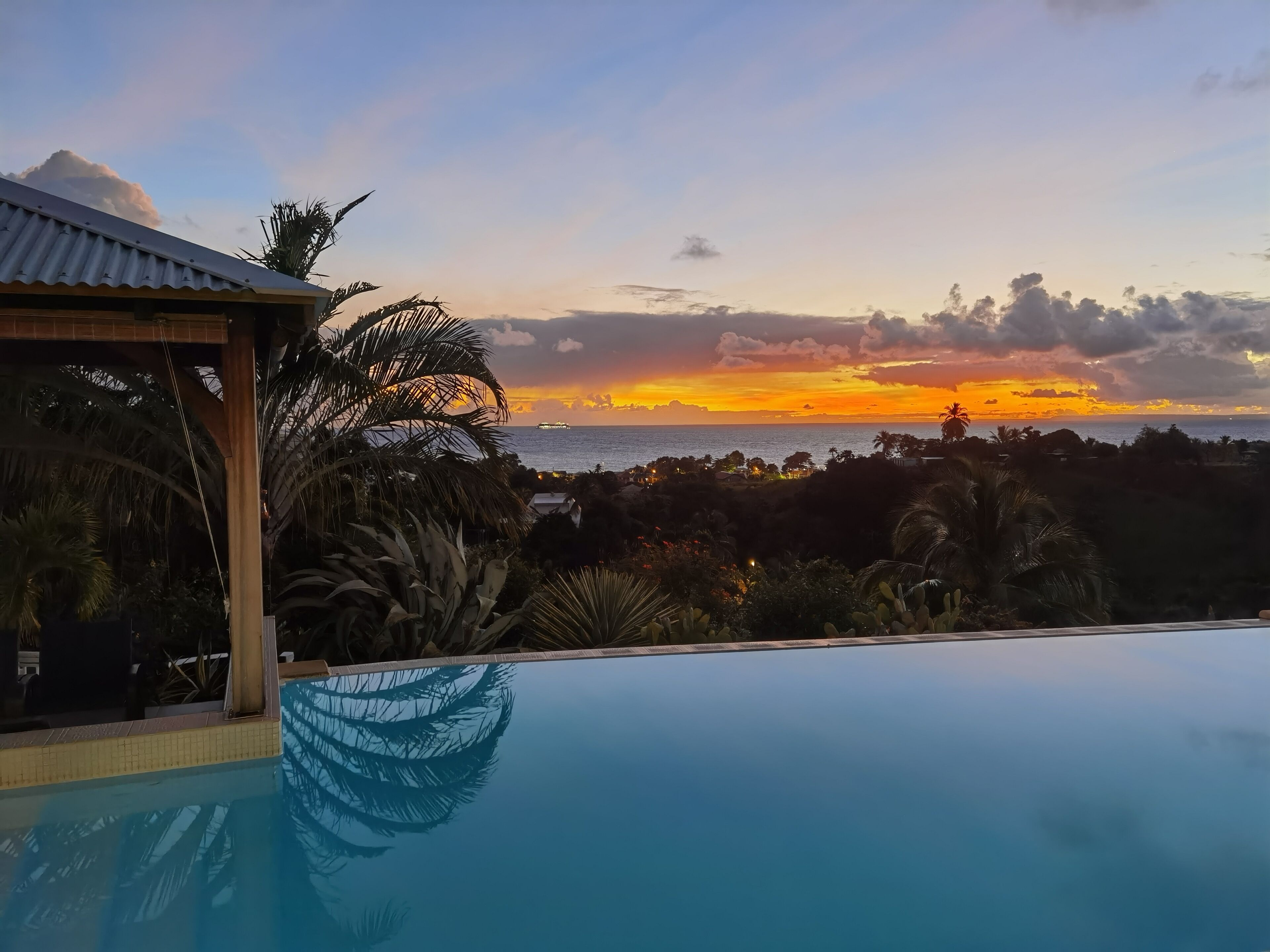 A pool with a view of a sunset over the ocean, surrounded by tropical vegetation.
