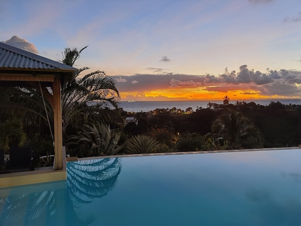 A pool with a view of a sunset over the ocean, surrounded by tropical vegetation.
