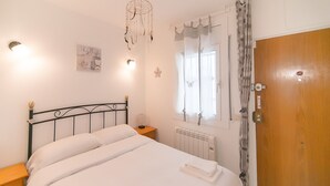 Een netjes opgemaakt bed in een kamer met een houten deur, een nachtkastje met een lamp en een raam met gordijnen.